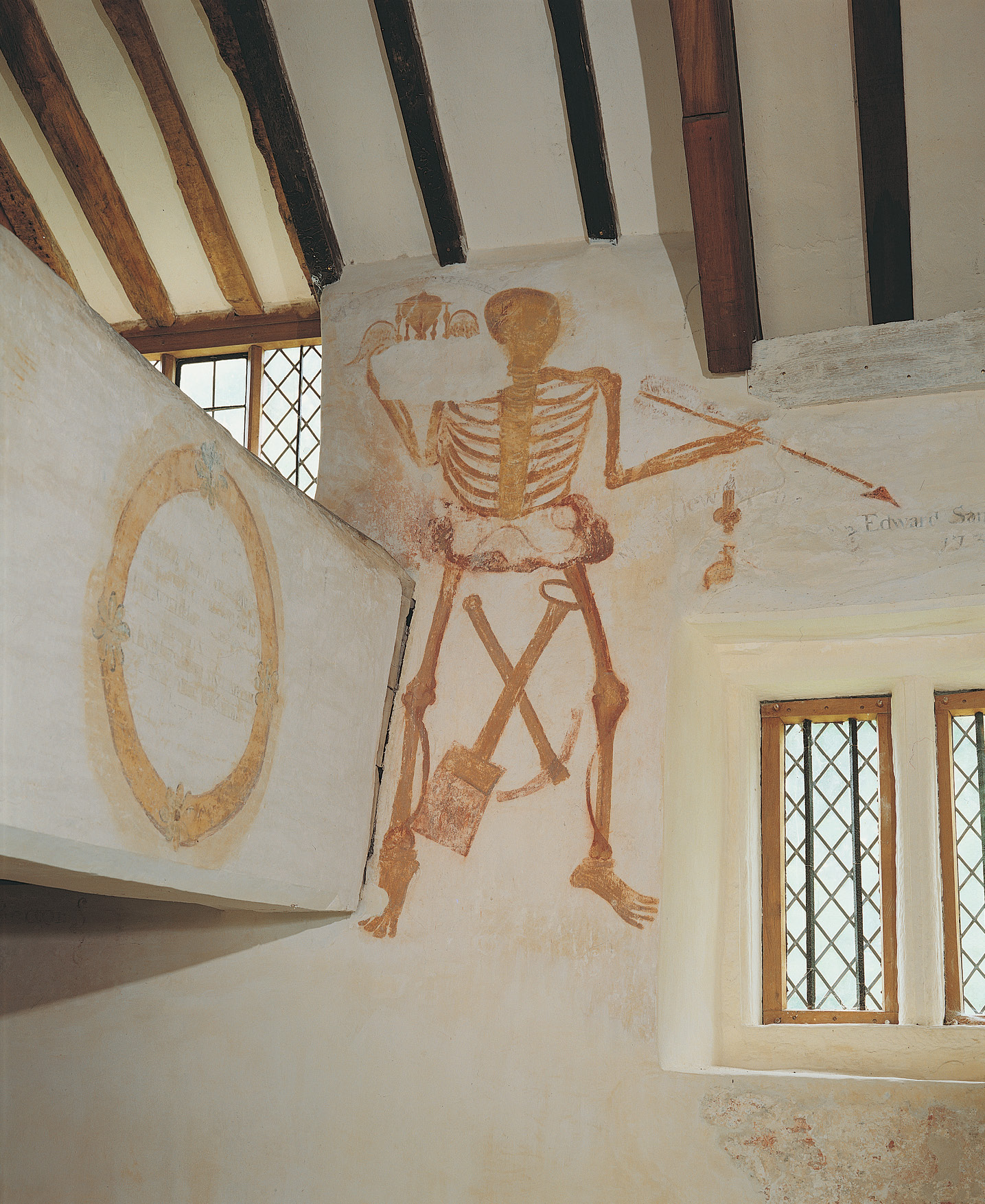 above A memento mori on the north wall of All Saints church, Llangar, Denbighshire, in the form of an arrow-wielding skeleton holding a winged hourglass, representing the speed with which a lifetime passes, and gravedigger’s mattock and spade.