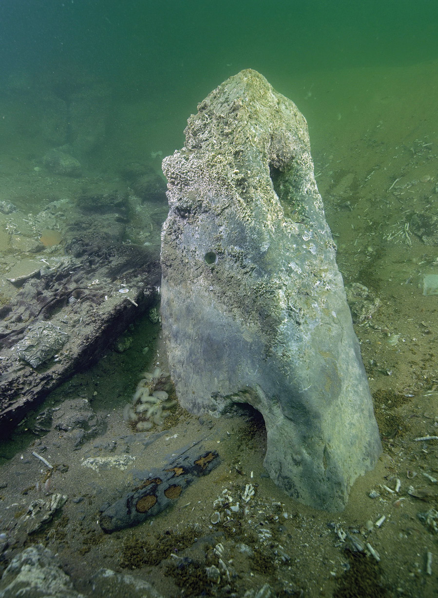 below A limestone anchor, with the hole at the top for rope cable that would have attached it to the ship, and a hole through the base where the remains of its wooden stake can still be seen.